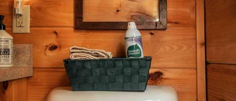 Un baño de madera con inodoro blanco, un cuadro de una hoja enmarco, una botella de jabón Seventh Generation y una cesta tejida con una toalla.
