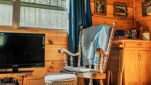 Interior acollidor d'una cabana amb elements de fusta, una televisió, una cadira amb una manta i una finestra amb cortines.