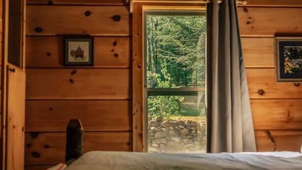 Habitació d'una cabana de fusta amb un llit, una finestra i una paret de pedra a l'exterior, i un quadre al mur.