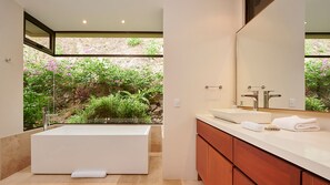 Une salle de bain moderne avec une grande baignoire, un lavabo et un miroir. La fenêtre offre une vue sur la verdure.