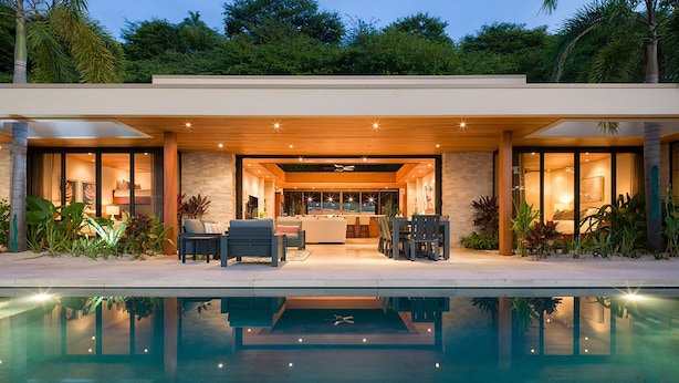 A modern house with a pool, outdoor seating, and a well-lit interior.