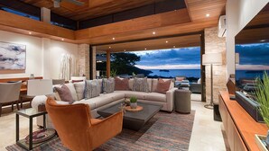 A modern living room with a large sectional sofa, a coffee table, and a view of the ocean at dusk.
