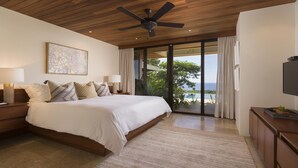 A bedroom with a large bed, a ceiling fan, a TV, and a view of the ocean through a sliding glass door.