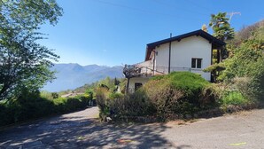 A two-story house with a balcony, surrounded by greenery and mountains in the background.