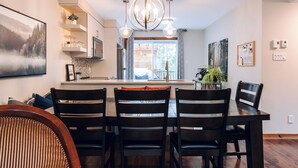 A dining area with a wooden table and chairs, a chandelier, and a view of the kitchen and outside.