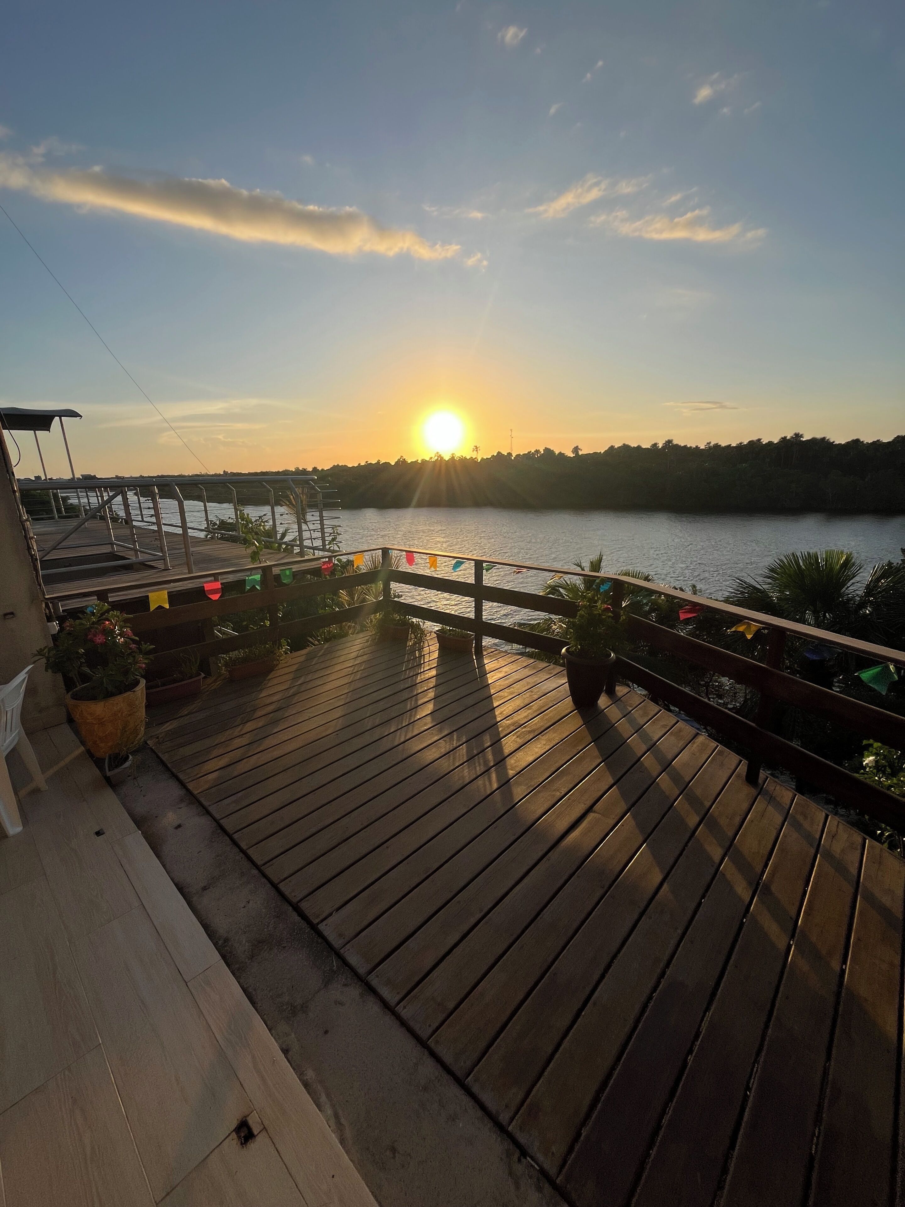 Une terrasse en bois agrémentée de plantes en pot, dotée d’une rambarde et offrant une vue sur une étendue d’eau au coucher du soleil.
