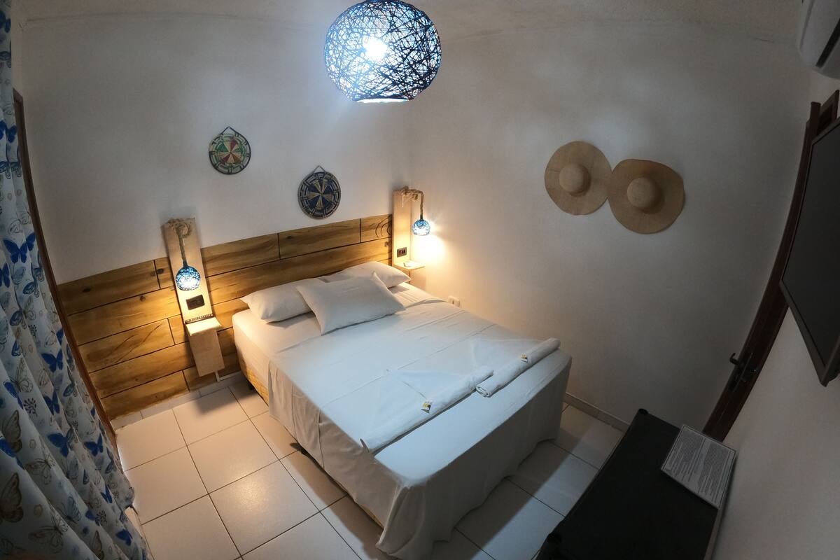 Cama bem arrumada com lençóis brancos, cabeceira de madeira, duas luminárias fixadas na parede e um ornamento decorativo na parede.