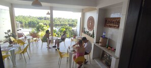 A dining area with yellow chairs and tables, a view of a river, and decorative wall art. - POUSADA VIDA NOVA BARREIRINHAS (Barreirinhas)