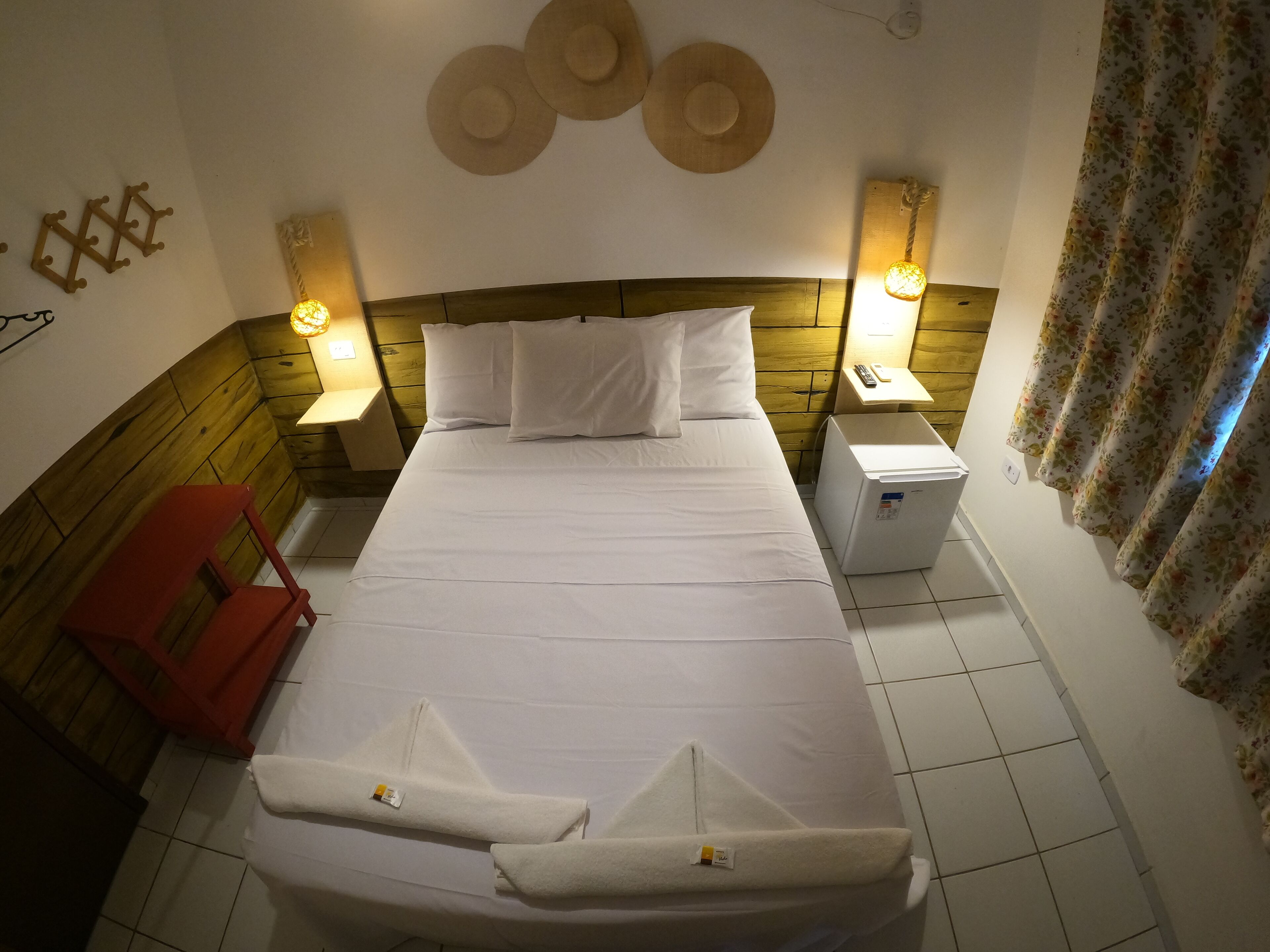 Una habitación de hotel con una cama, dos mesitas de noche, una silla roja, un perchero en la pared y un refrigerador pequeño.