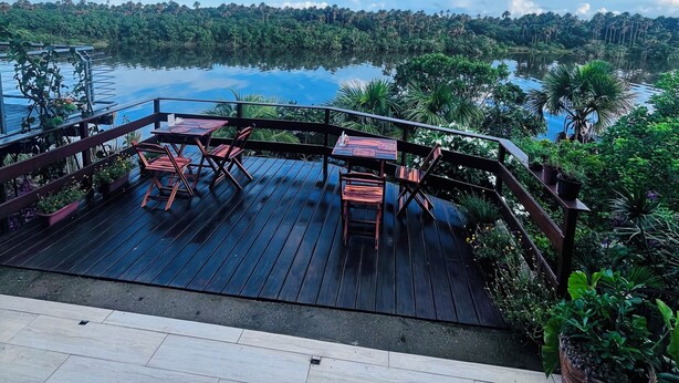 Una terraza de madera con mobiliario exterior, barandilla y vistas a un lago y un bosque.