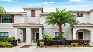 A multi-story residential building with a tiled roof, a central entrance with a covered porch, and a well-maintained garden with palm trees and shrubs.