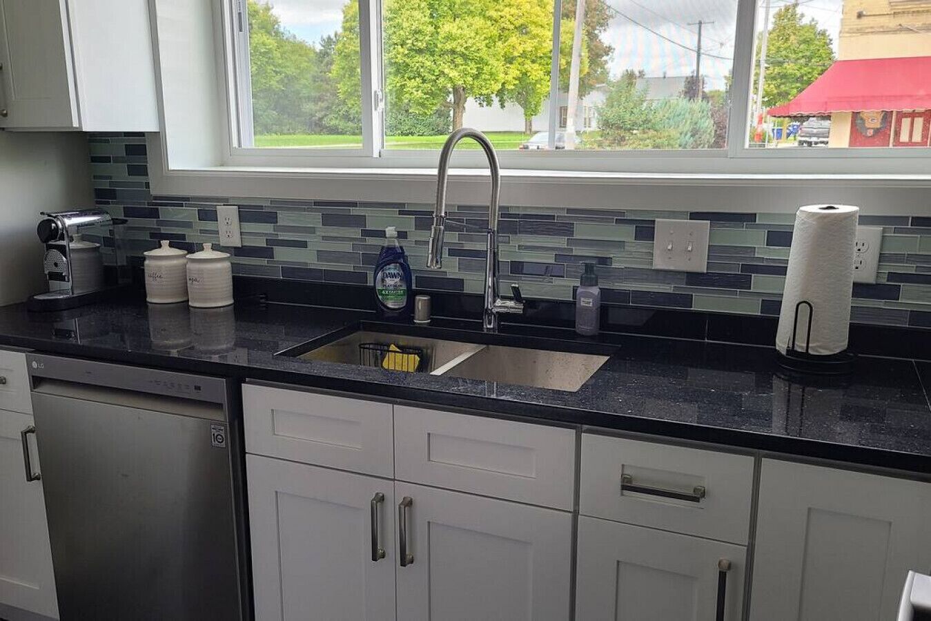 A kitchen with a black countertop, white cabinets, a stainless steel sink, and a window with a view of trees and buildings.