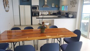 A modern kitchen with white cabinets, a built-in oven, and a microwave. There is a dining area with a wooden table and blue chairs.