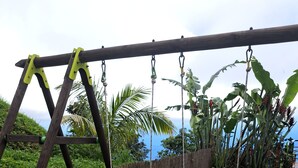 A wooden swing set with two swings, a green slide, and a small table with chairs in a backyard with artificial grass and tropical plants.