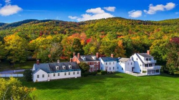 A cluster of white buildings with multiple chimneys, surrounded by lush green grass and trees with autumn foliage.