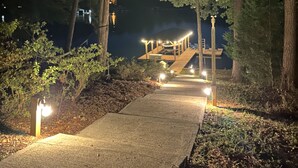 Un sendero nocturno que conduce a una estructura de madera con techo, rodeada de árboles e iluminada por luces en el suelo.