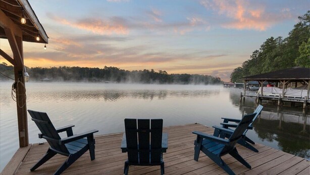 Una terraza de madera con dos sillas Adirondack mirando un lago tranquilo al atardecer.