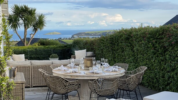 An outdoor dining area with wicker furniture, a round table set for six, and a view of the sea and hills in the background. - Trem Ynys (Pwllheli)