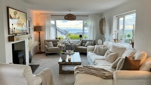A spacious living room with a fireplace, two sofas, a coffee table, and large windows offering a view of the sea. - Trem Ynys (Pwllheli)