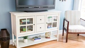 A white entertainment center with a flat-screen TV, glass-fronted cabinets, and a striped chair. - The City Cottage, pet friendly, walk to shops, food & NCSU (Raleigh)