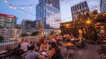 An outdoor dining area with tables and chairs, surrounded by city buildings and string lights.