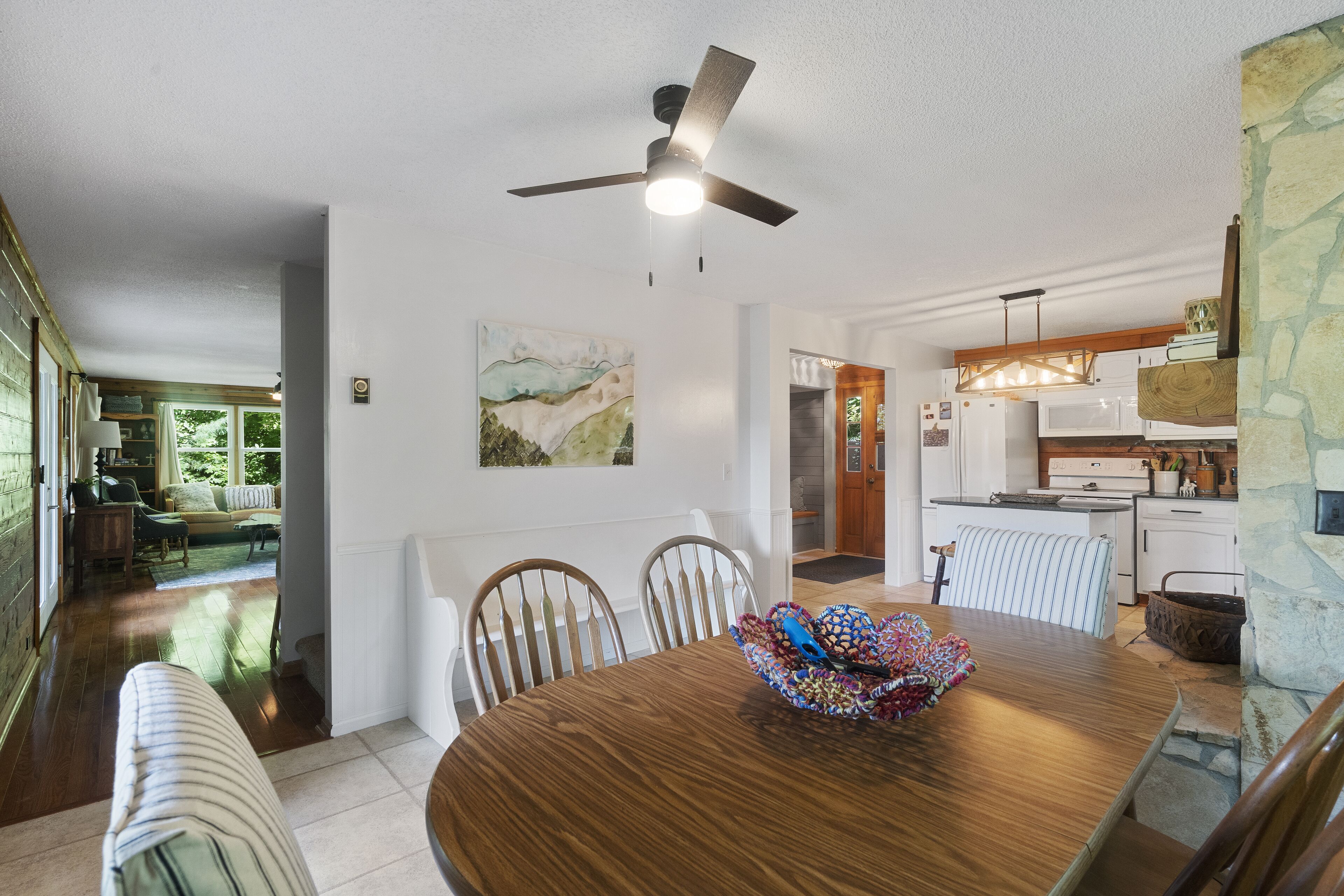 A dining area with a wooden table, chairs, and a ceiling fan. A kitchen is visible in the background with white cabinets and a stone accent wall.