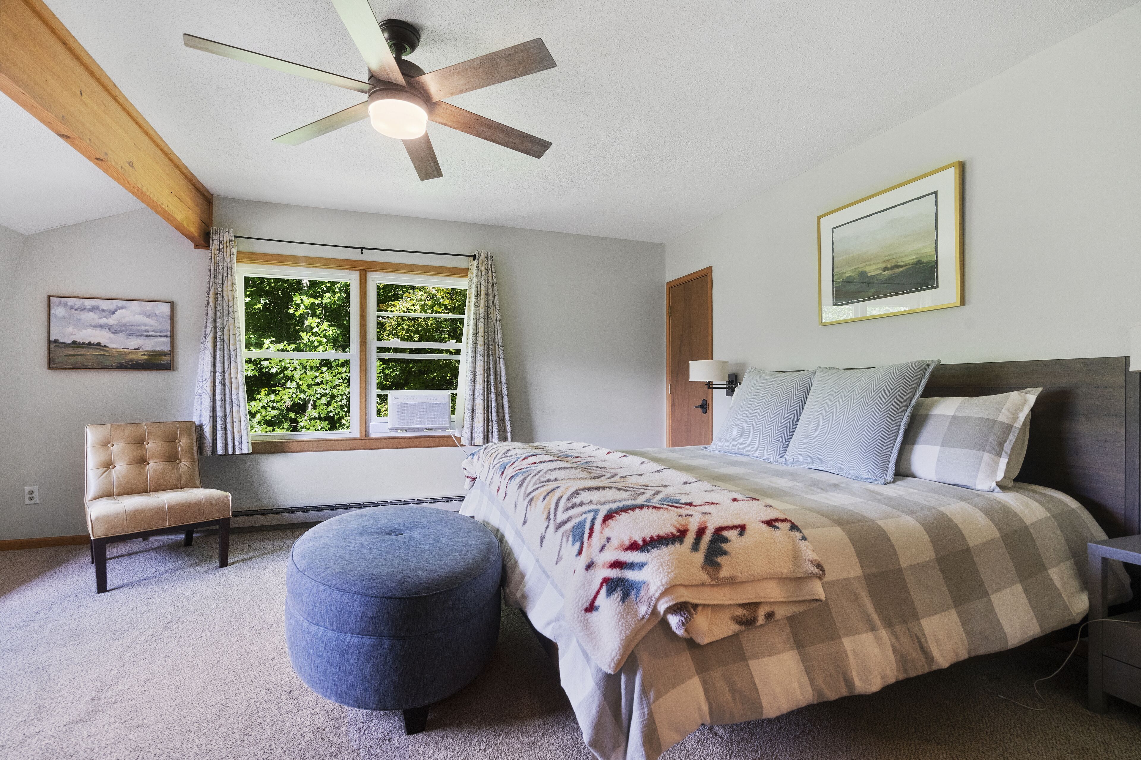 A bedroom with a bed, a chair, a round ottoman, a ceiling fan, a window with curtains, and a framed picture on the wall.