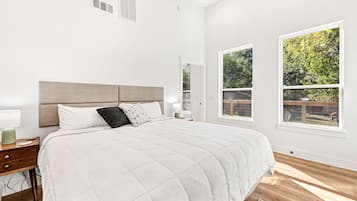 A bedroom with a large bed, a nightstand, a lamp, and two windows with a view of greenery.