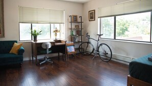 Una habitación con suelo de madera, un sofá verde, un escritorio con una silla, una bicicleta y ventanas grandes con persianas.