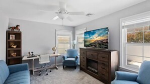 Una habitación con escritorio, silla, televisión, estantería y dos sofás.
