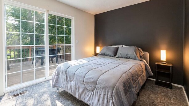 Une chambre avec une grande porte-fenêtre coulissante donnant sur une terrasse, un lit avec une couette grise, deux lampes de chevet et un mur d’accent de couleur foncée.