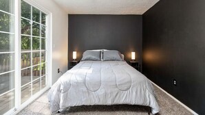 Une chambre avec une grande fenêtre, un lit, deux tables de chevet avec des lampes et un mur de couleur sombre.