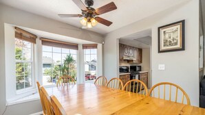 Une salle à manger avec une table et des chaises en bois, un ventilateur de plafond et de grandes fenêtres équipées de stores.