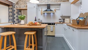 Una cocina con pared de piedra, vigas de madera, una isla grande con taburetes, una lámpara colgante y una cesta de aperitivos.