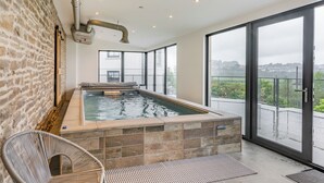 Una piscina interior moderna con paredes de piedra, una puerta de vidrio que da al exterior y una silla de mimbre.