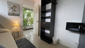 A modern hotel room with a bed, a bedside table with a lamp, a black shelving unit, a white trash bin, and a door leading to a balcony with a view of trees. - Escape a la Ciudad en Nuestro Encantador Apartamento con Todas las Comodidades (San Rafael Arriba de Desamparados)
