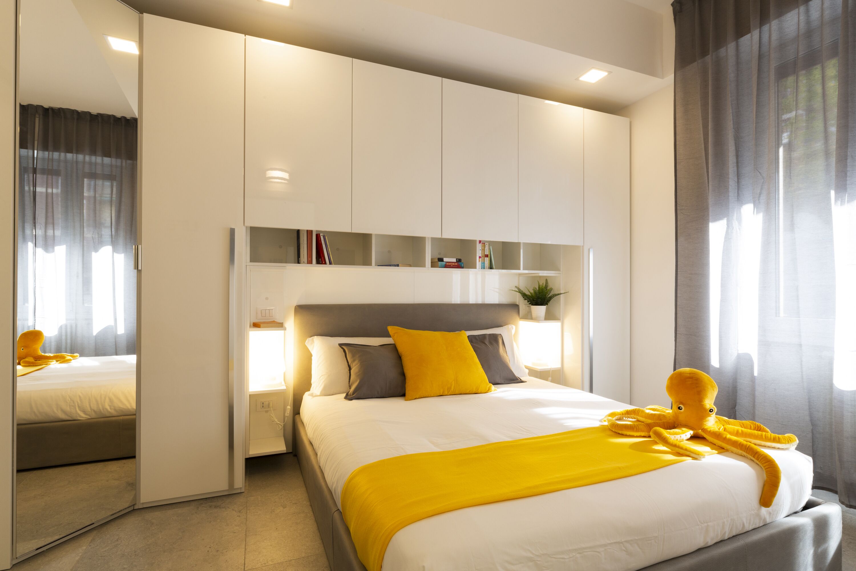 A modern hotel room with a large bed, a bedside table with a lamp, a mirror, and a wardrobe with built-in shelves.