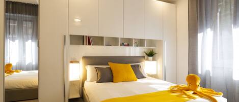 A modern hotel room with a large bed, a bedside table with a lamp, a mirror, and a wardrobe with built-in shelves.