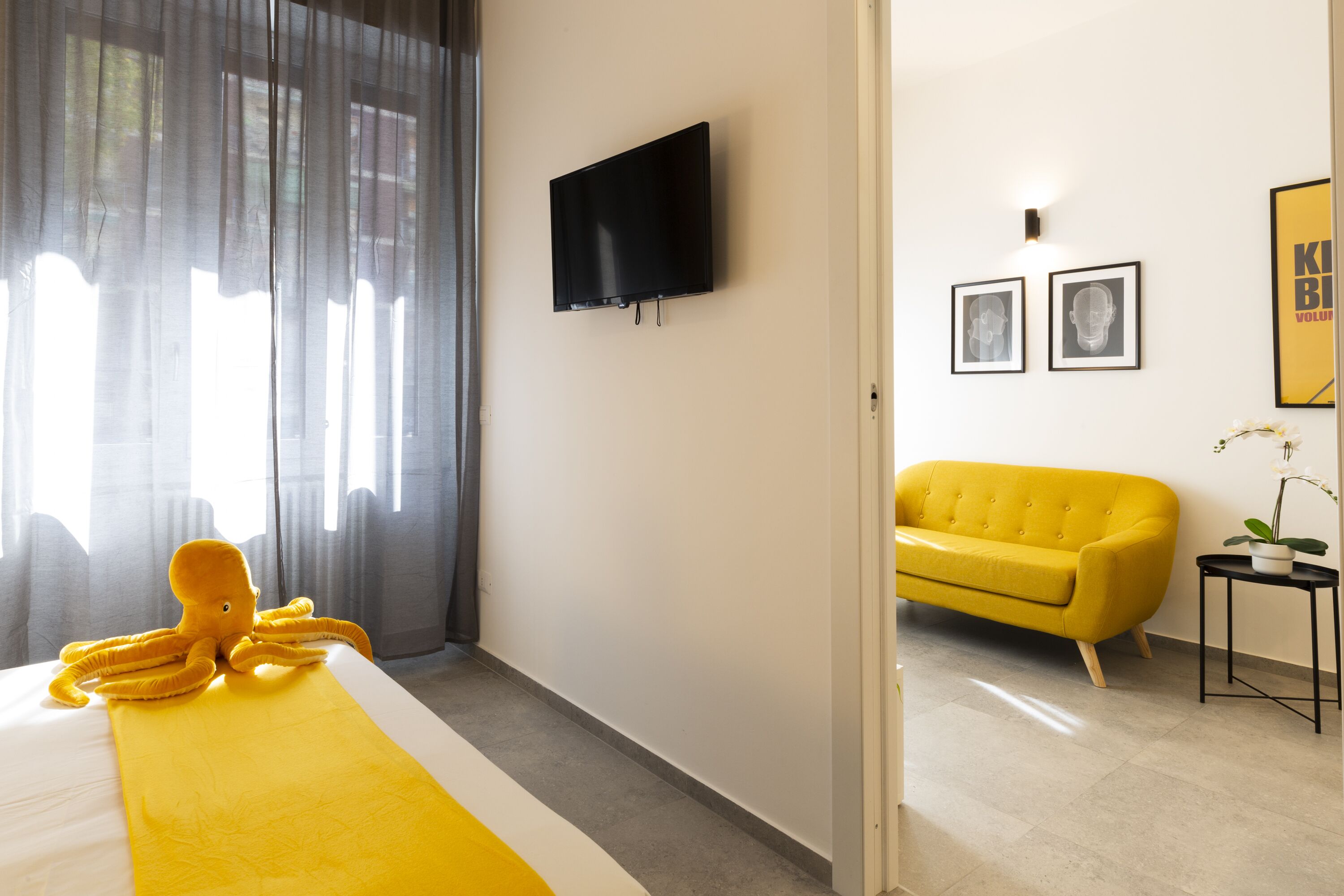 Habitación de hotel con una cama, un televisor montado en la pared, un sofá amarillo y una pequeña mesa con una planta.
