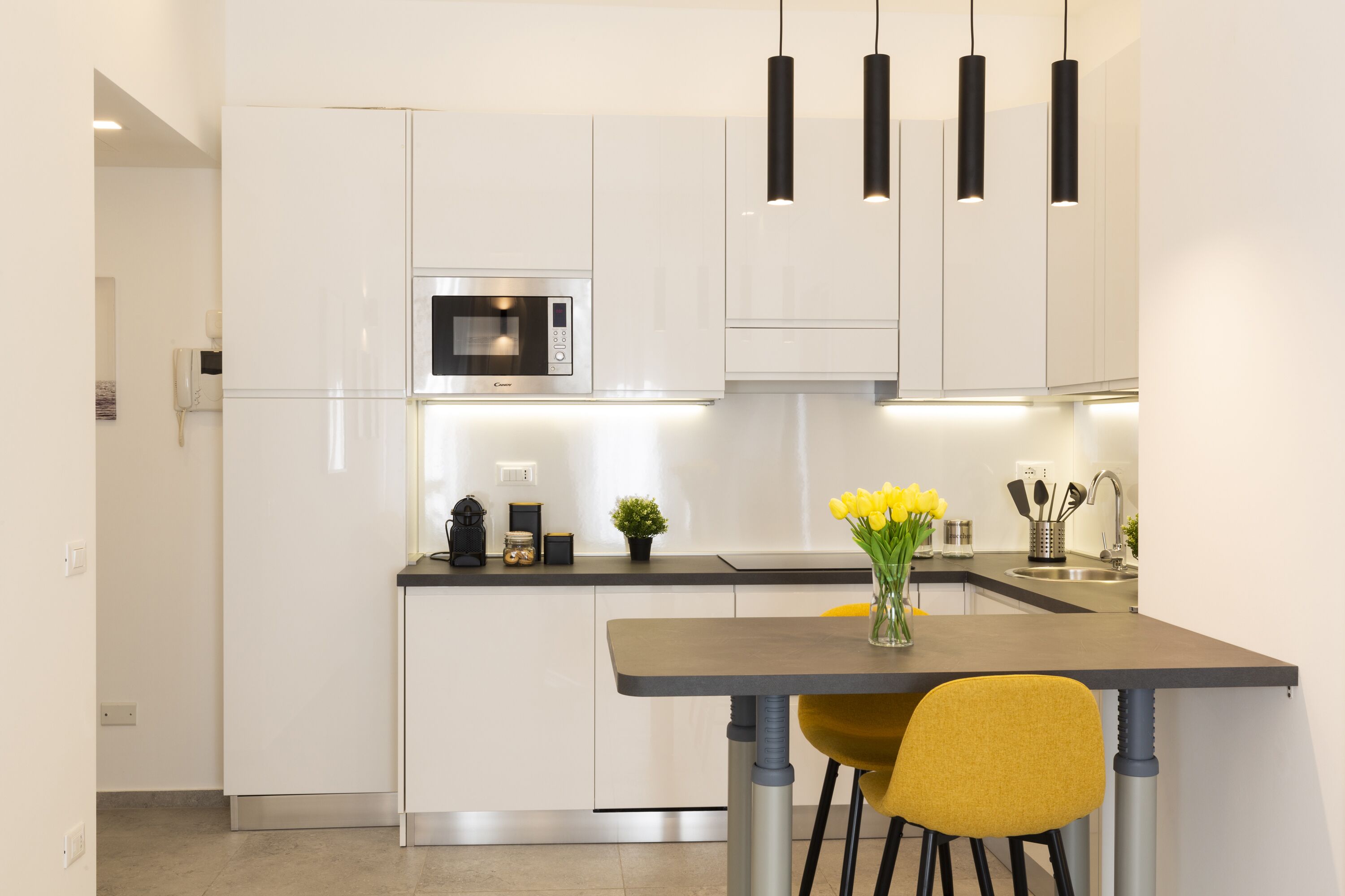 Una cocina moderna con armarios blancos, microondas integrado, una pequeña isla con un florero de flores amarillas y lámparas colgantes negras.