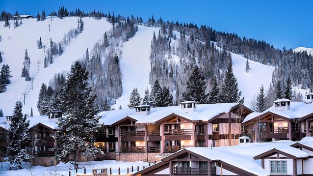 Un chalet enneigé composé de plusieurs bâtiments, avec une piste de ski en arrière-plan et une montagne boisée.