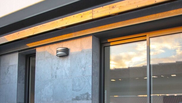 Un bâtiment moderne avec une façade en pierre, de grandes portes vitrées et une poutre en bois au-dessus de l’entrée.