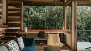 A modern interior with a wooden ceiling, a sofa with patterned cushions, a desk with a chair, and large windows offering a view of trees.