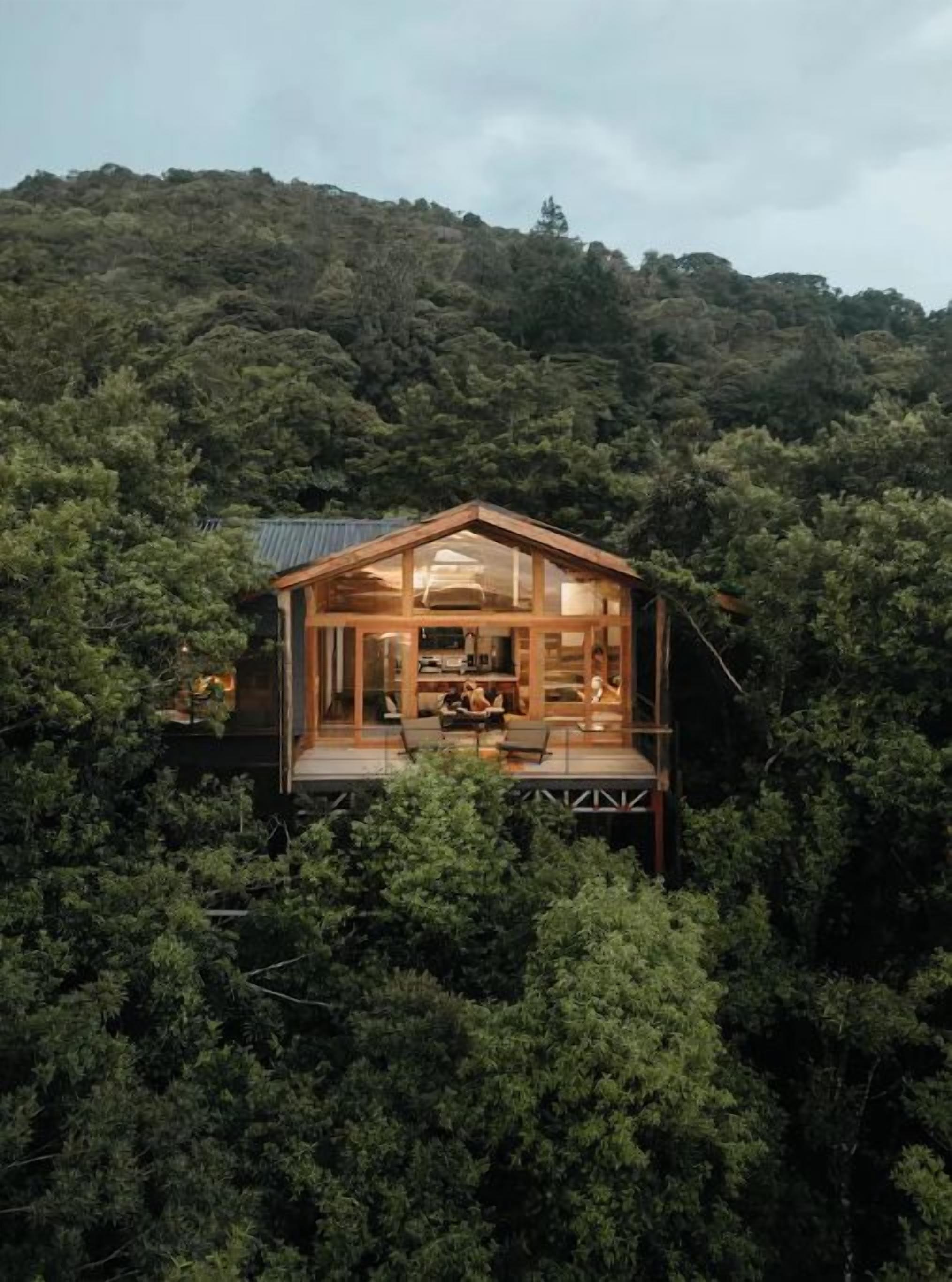 Una cabaña de madera con fachada de vidrio, rodeada por un bosque denso, que cuenta con una terraza y un interior visible con zona de comedor.