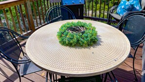 A round outdoor table with a woven edge, surrounded by metal chairs, on a wooden deck with a grill and chairs in the background.