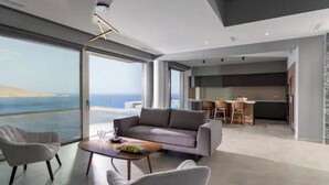 A modern living room with a sofa and chairs, a dining area with a table, and a kitchen in the background. - Luxury Seafront Villa Eros with Private Pool - Rock Bay Villas - Crete, Greece (Agia Pelagia)