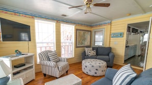 A cozy living room with a gray sofa, striped armchair, and ottoman, a television mounted on the wall, and a ceiling fan. - Historic 1940's Beach Cottage, steps to the beach!   (Tybee Island)