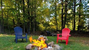 A campfire with two Adirondack chairs, one blue and one red, in a wooded area. - Cozy Adirondack Retreat sleeps 5, 3 miles to Gore skiing, hiking, rafting (North Creek)