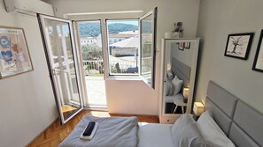 A bedroom with a bed, a bedside table, a nightstand, a window with a view of buildings and hills, and a wall-mounted shelf with a plant. - Oleas Stari Grad Hvar – Holiday Home – 2 min to Sea and Center (Stari Grad)