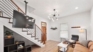 A modern living room with a staircase, a flat-screen TV, a chandelier, a sofa, and a coffee table.
